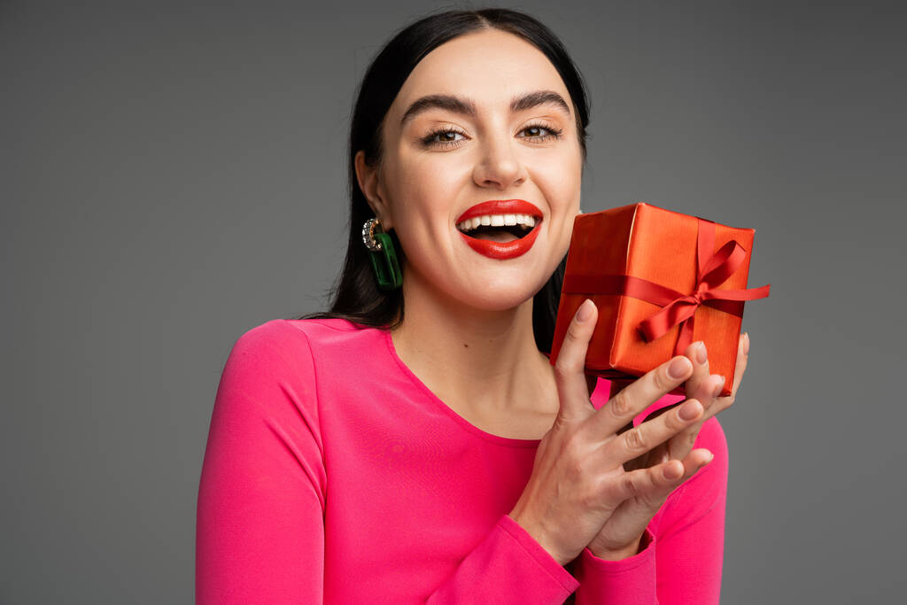 opgewonden en chique jonge vrouw met trendy oorbellen en vlekkeloze make-up glimlachen terwijl het houden van rode en gewikkelde geschenkdoos op grijze achtergrond  - Foto, afbeelding