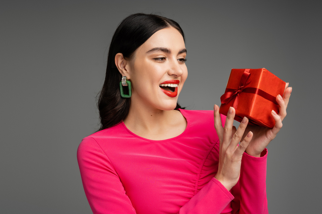 izgatott és elegáns fiatal nő divatos fülbevalók és fényes barna haj mosolyog, miközben gazdaság piros és csomagolt ajándék doboz szürke háttér  - Fotó, kép