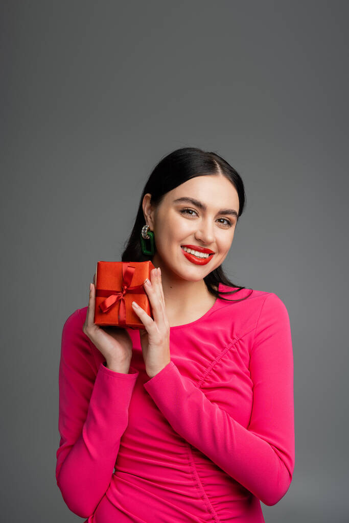 elegant and young woman with trendy earrings and brunette hair smiling while holding red and wrapped present for holiday on grey background  - Photo, Image