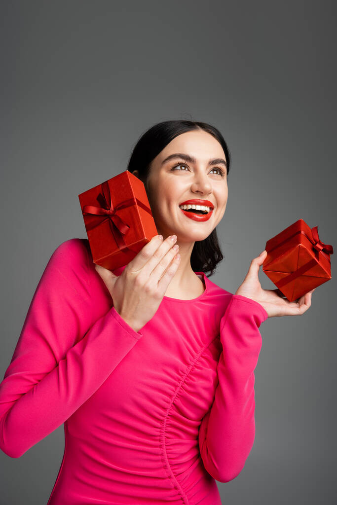 χαρούμενη νεαρή γυναίκα με καστανά μαλλιά και μοντέρνα σκουλαρίκια χαμογελώντας ενώ στέκεται σε μωβ φόρεμα κόμμα και κρατώντας τυλιγμένο κουτιά δώρων για διακοπές σε γκρι φόντο  - Φωτογραφία, εικόνα