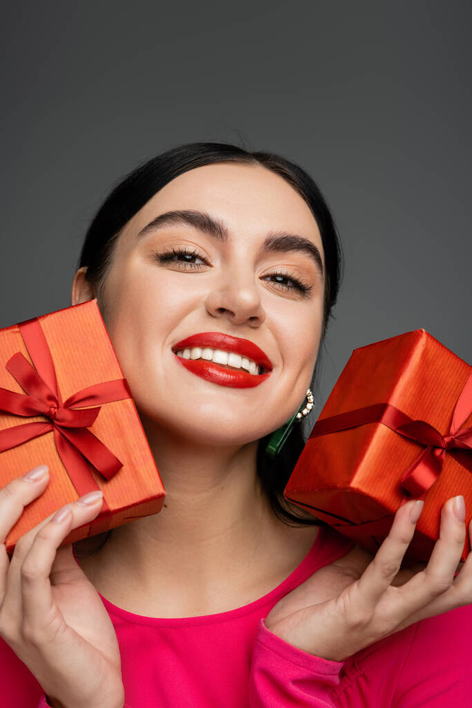 portrait of cheerful young woman with shiny brunette hair and trendy earrings smiling while holding wrapped gift boxes for holiday and looking at camera on grey background  - Photo, Image