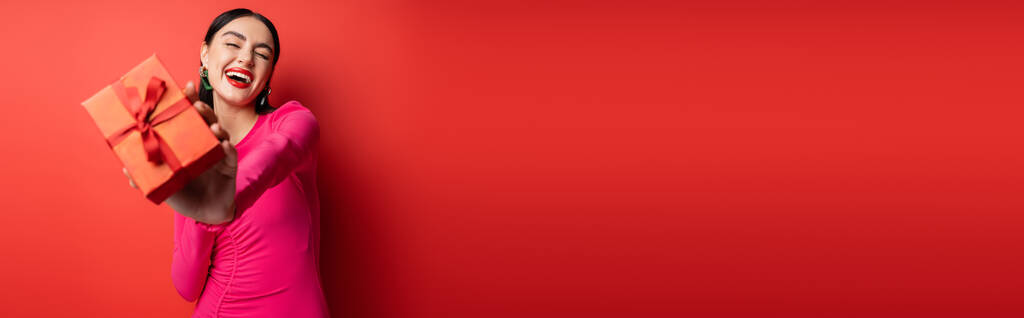 urocza kobieta z brunetką włosy i modne kolczyki uśmiechnięta stojąc w sukni magenta party i trzymając zapakowany prezent na wakacje na czerwonym tle, baner  - Zdjęcie, obraz