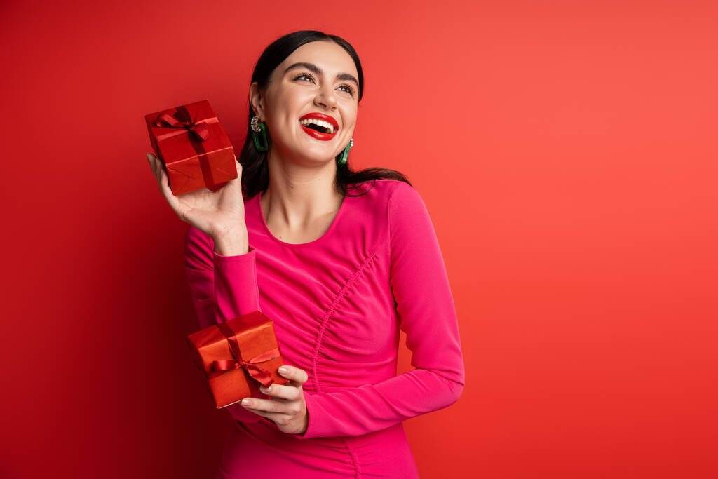 donna felicissima con capelli castani e orecchini alla moda sorridenti mentre in piedi in abito da festa magenta e con in mano scatole regalo avvolte per le vacanze su sfondo rosso  - Foto, immagini