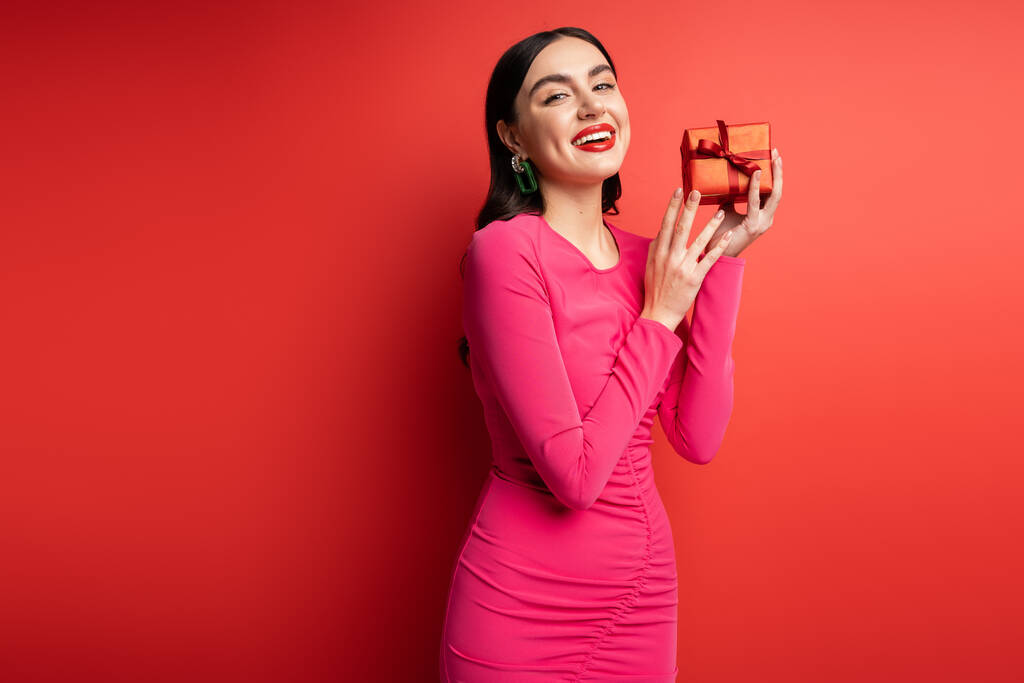 λαμπερό γυναίκα με καστανά μαλλιά και μοντέρνα σκουλαρίκια χαμογελώντας, ενώ στέκεται σε μωβ φόρεμα κόμμα και κρατώντας τυλιγμένο δώρο έκπληξη για διακοπές σε κόκκινο φόντο  - Φωτογραφία, εικόνα