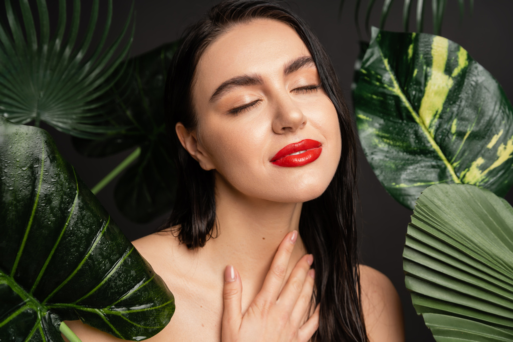 gorgeous young woman with brunette hair and red lips smiling while posing with closed eyes around tropical, wet and green palm leaves with raindrops on them   - Photo, Image