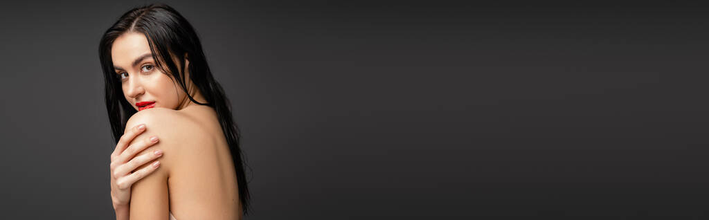 ritratto di giovane e bella donna con capelli bagnati e bruni che toccano la spalla nuda mentre guarda la fotocamera dopo la doccia isolata su sfondo grigio, banner  - Foto, immagini