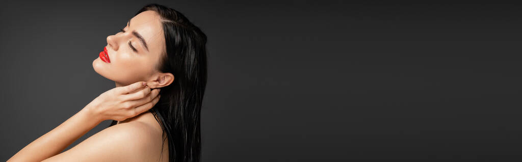 hermosa y joven mujer con el pelo moreno húmedo y labios rojos tocando el cuello mientras posa con los hombros desnudos después de la ducha aislado sobre fondo gris, pancarta  - Foto, imagen
