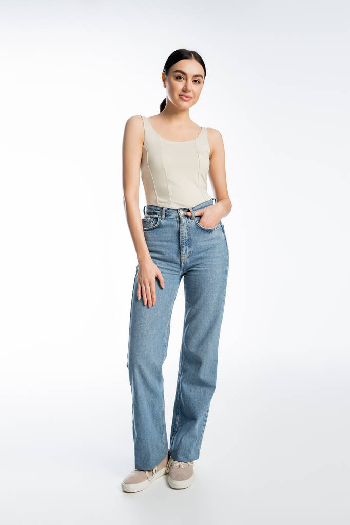 full length of charming young brunette woman standing in blue denim jeans and tank top while posing with hand in pocket and looking at camera on white background  - Photo, Image