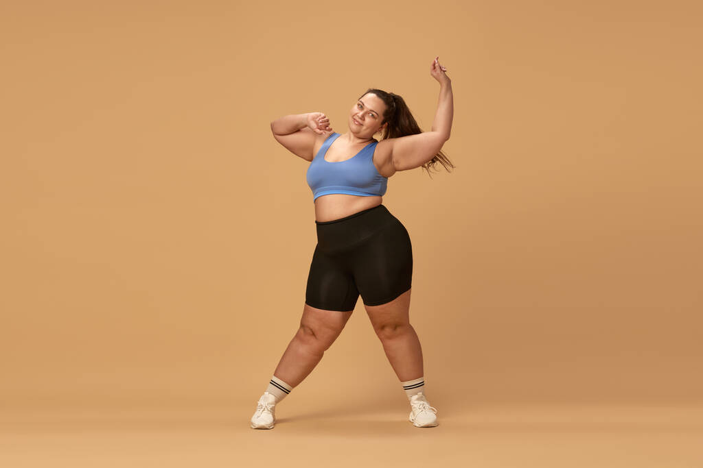 Молодая толстая женщина тренируется в спортивной одежде на фоне коричневой студии. Потеря веса с фитнесом и диетой. Концепция спорта, телосложения, похудения, ухода за телом и здоровьем - Фото, изображение