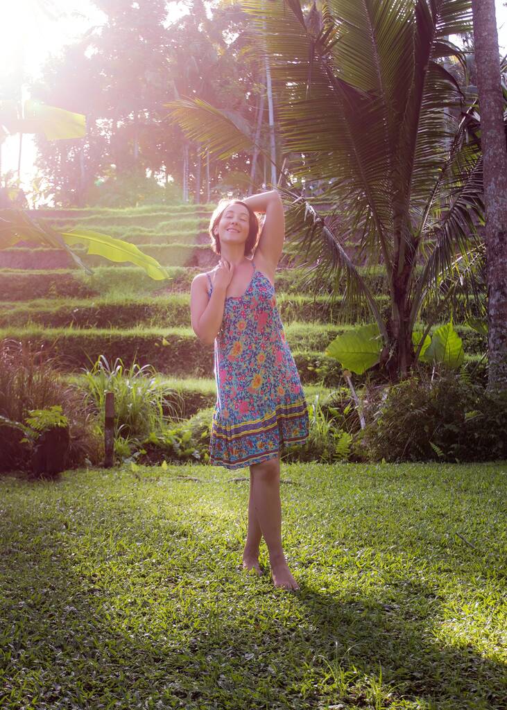 Pystysuora kuva nuoresta naisesta kesämekossa nauttimassa aamuvalosta trooppisessa puutarhassa. - Valokuva, kuva