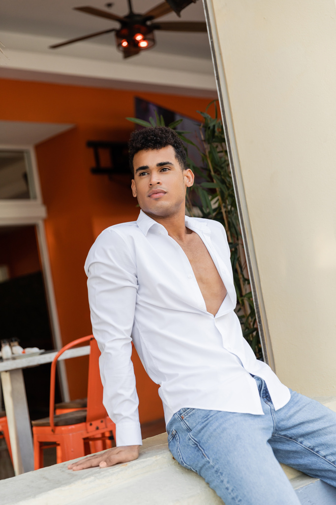 hombre cubano rizado en camisa blanca y jeans sentados en parapeto de café al aire libre con palmeras en Miami - Foto, Imagen