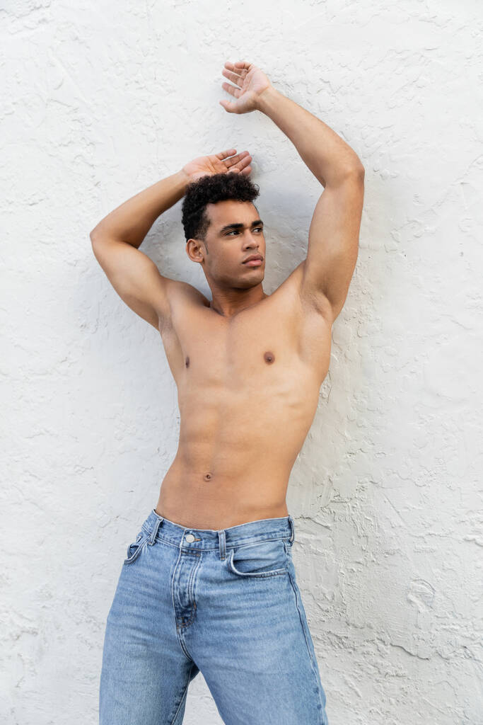 Muscoloso e di bell'aspetto giovane cubano in jeans blu in posa vicino al muro bianco in strada a Miami - Foto, immagini