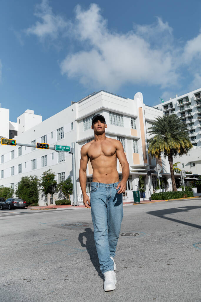 hemdloser junger kubanischer Mann in Jeans und Baseballkappe, der im Sommer auf der städtischen Straße in Miam spaziert  - Foto, Bild