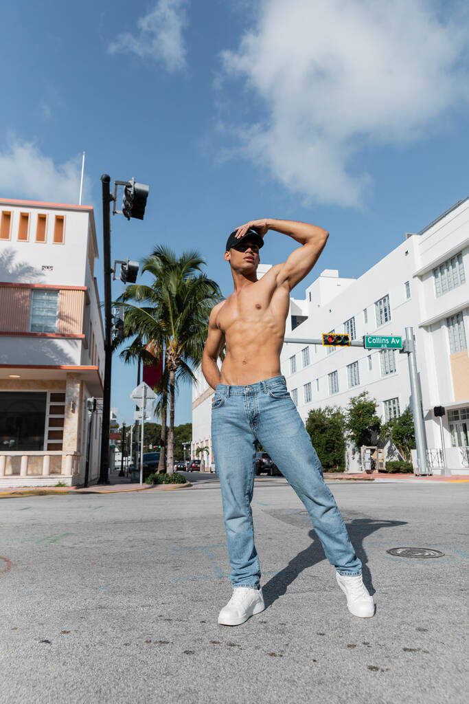 horký kubánský muž s atletickým tělem v baseballové čepici a modrými džínami na ulici v Miami, jižní pláž - Fotografie, Obrázek