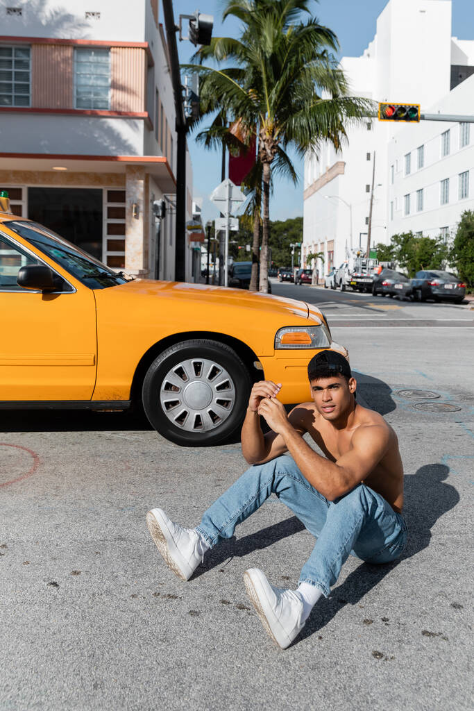 Sexy cuban man with athletic body in baseball cap sitting on road near yellow taxi in Miami - Photo, Image