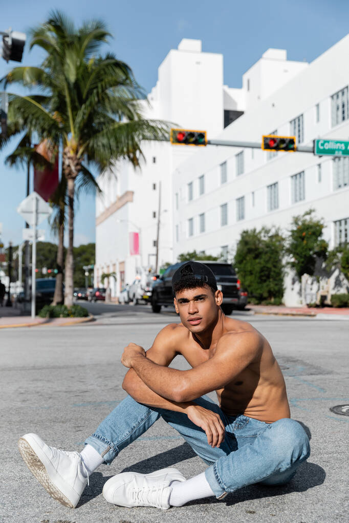 homme cubain sexy en casquette de baseball et jeans bleus assis sur la route à Miami  - Photo, image