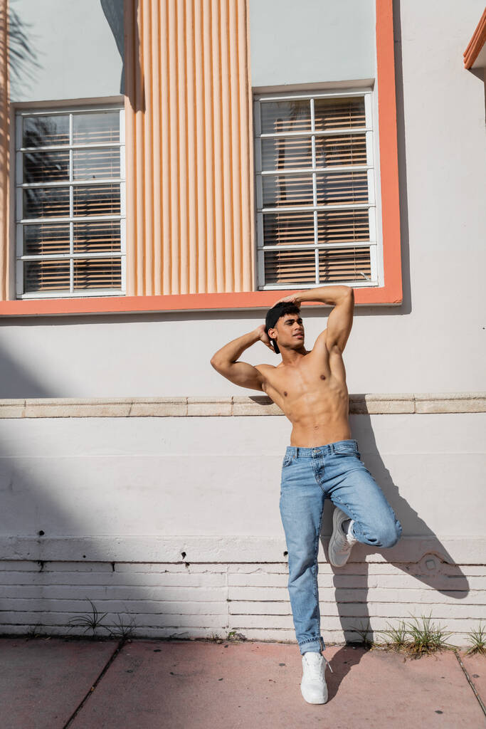 Sexy muskulöser kubanischer Mann mit athletischem Körper in Basecap und Jeans steht auf der Straße in Miami - Foto, Bild