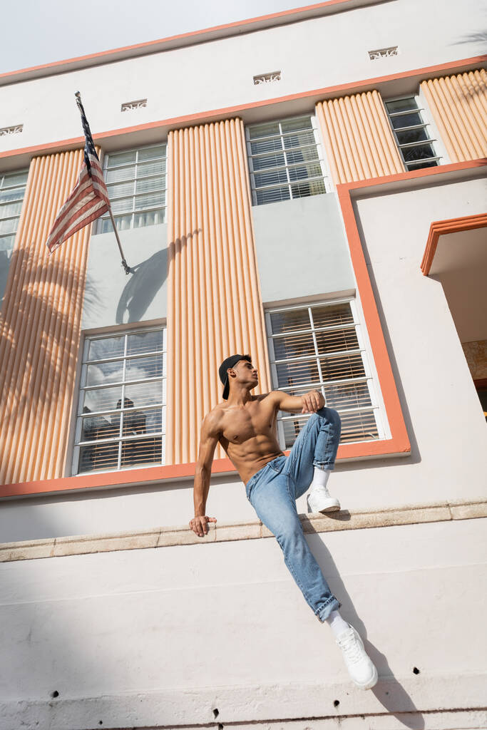 Kubańczyk z muskularnym ciałem pozujący latem w czapce baseballowej i dżinsach na ulicy w Miami - Zdjęcie, obraz
