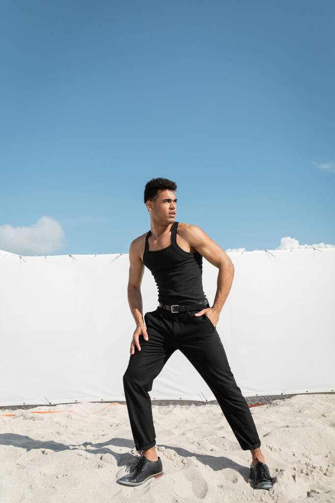 Przystojny Kubańczyk w czarnej koszulce bez rękawów i spodniach stojący na piasku w Miami, na południowej plaży - Zdjęcie, obraz