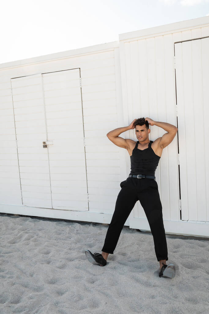 Relaxed and muscular young cuban man in black outfit touching head in Miami, south beach - Photo, Image