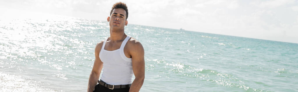 elegante giovane cubano guardando la fotocamera con acqua dell'oceano e cielo sullo sfondo a Miami, banner  - Foto, immagini