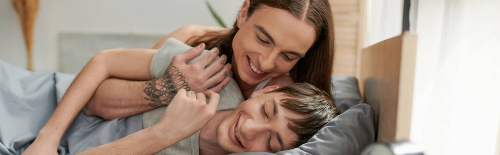 Τατουάζ και μακριά μαλλιά γκέι άντρας αγκαλιάζει χαρούμενο φίλο με κλειστά μάτια, ενώ ξυπνάει στο άνετο κρεβάτι το πρωί, πανό  - Φωτογραφία, εικόνα