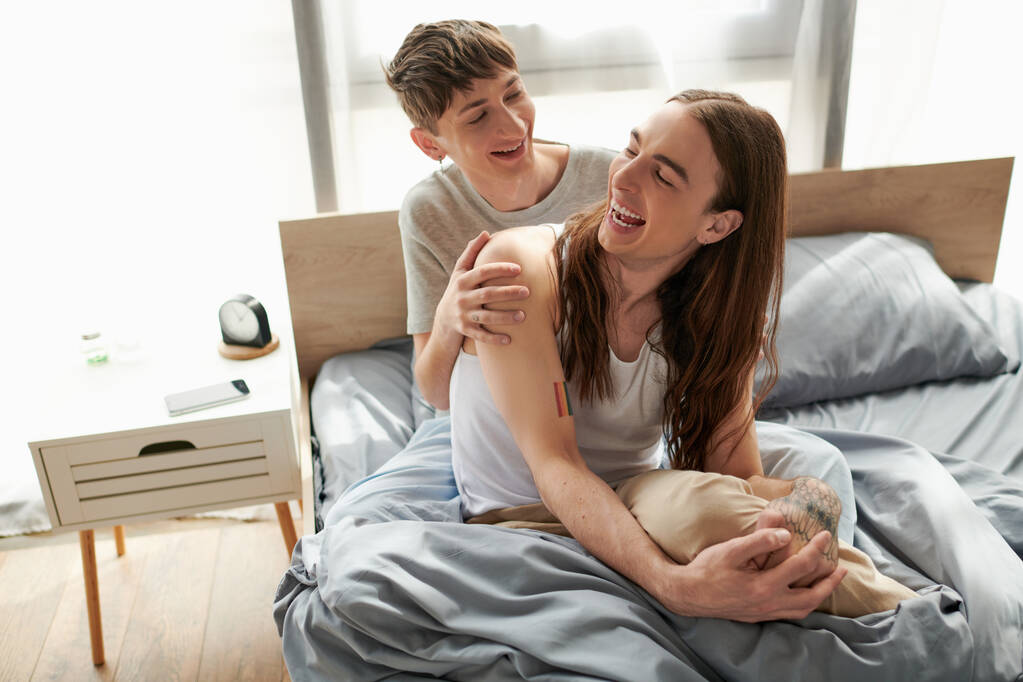 Iloinen ja nuori homopari pyjamassa viettää aikaa yhdessä ja puhuu toisilleen lepäämällä mukavalla sängyllä herättyään aamulla kotona.  - Valokuva, kuva