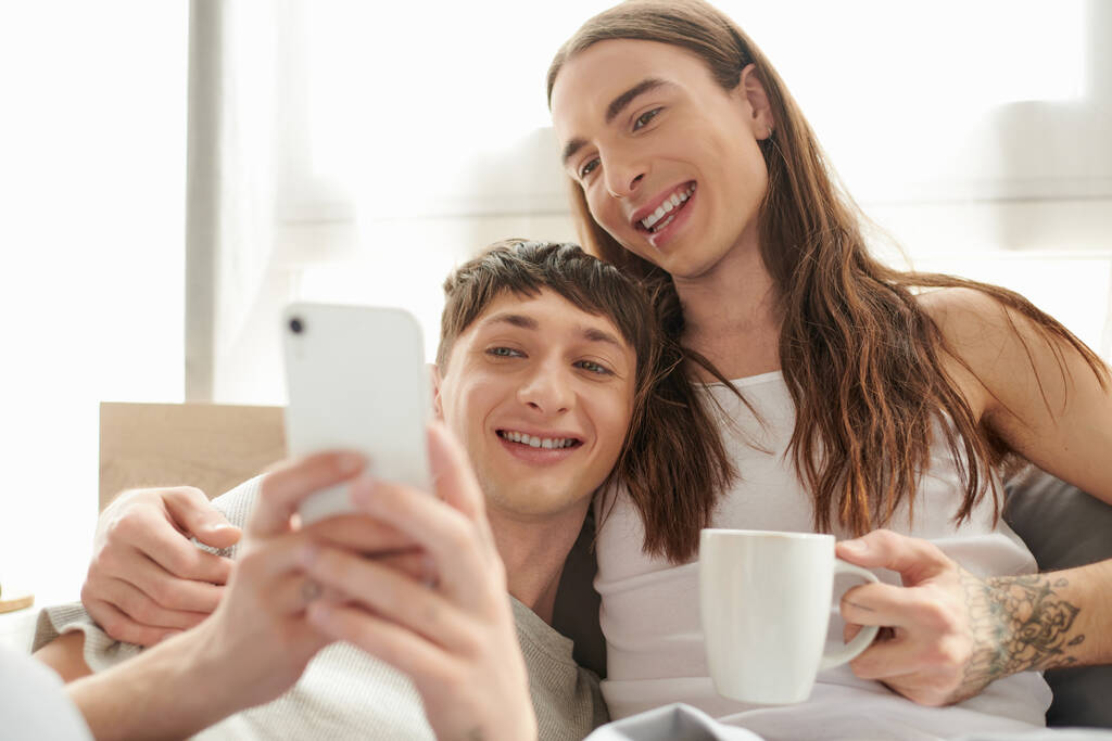 Young same sex couple in pajama holding coffee cup and smiling while using blurred smartphone and resting on comfortable bed in bedroom in morning at home  - Photo, Image