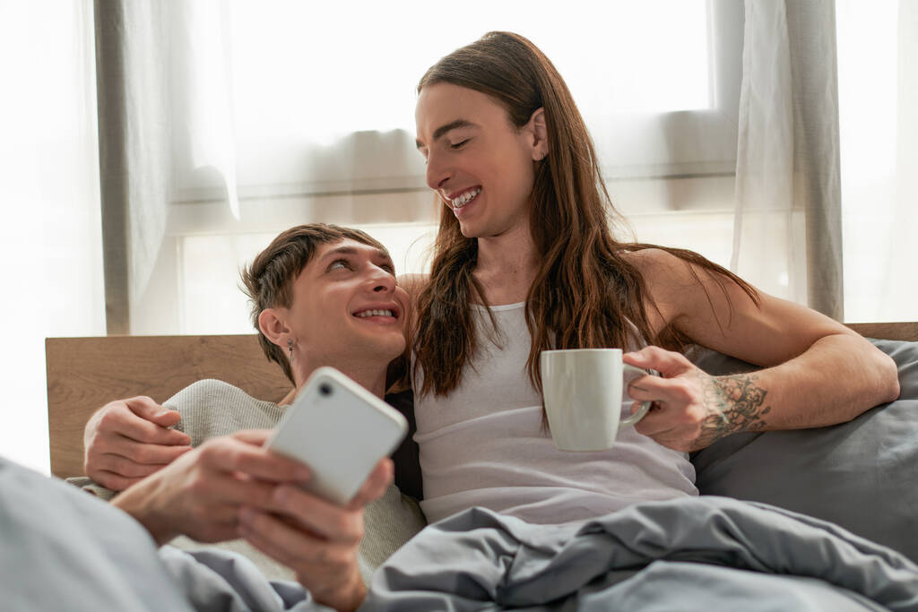 Positive homosexual man holding coffee cup while hugging and looking at boyfriend in pajama using blurred smartphone on comfortable bed at home in morning  - Photo, Image