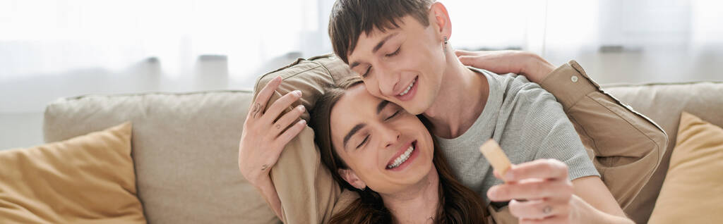 Joven y positiva pareja del mismo sexo cerrando los ojos mientras abraza y juega bloques de madera juego cerca de sofá en la sala de estar en casa, pancarta  - Foto, imagen