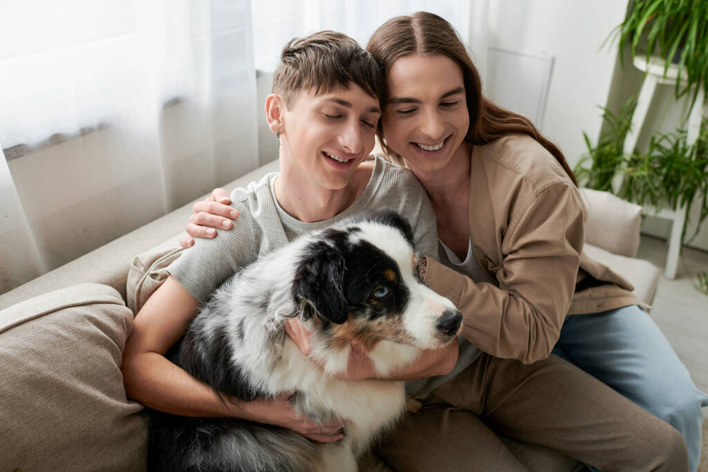 High angle view of smiling homosexual couple with closed eyes hugging near Australian shepherd dog on couch in modern living room at home  - Photo, Image