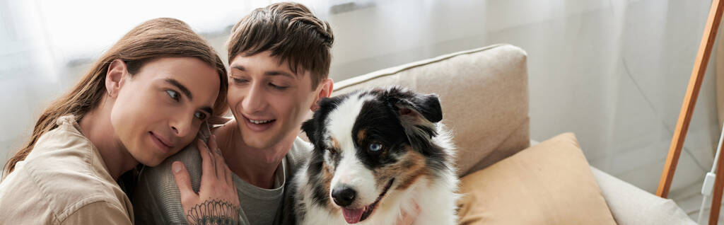 Hosszú hajú meleg férfi tetoválással a kezén ölelés mosolygós barátja alkalmi ruhák közelében ausztrál pásztor kutya modern kanapén a nappaliban otthon, banner  - Fotó, kép