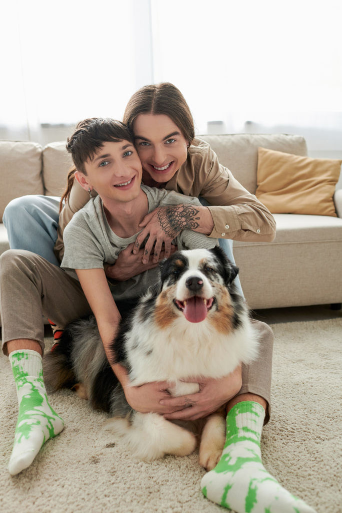 Vrolijk en jong lgbt paar in casual kleding en sokken knuffelen en kijken naar de camera in de buurt van harige Australische herder hond liggend op tapijt op de vloer in de moderne woonkamer thuis  - Foto, afbeelding