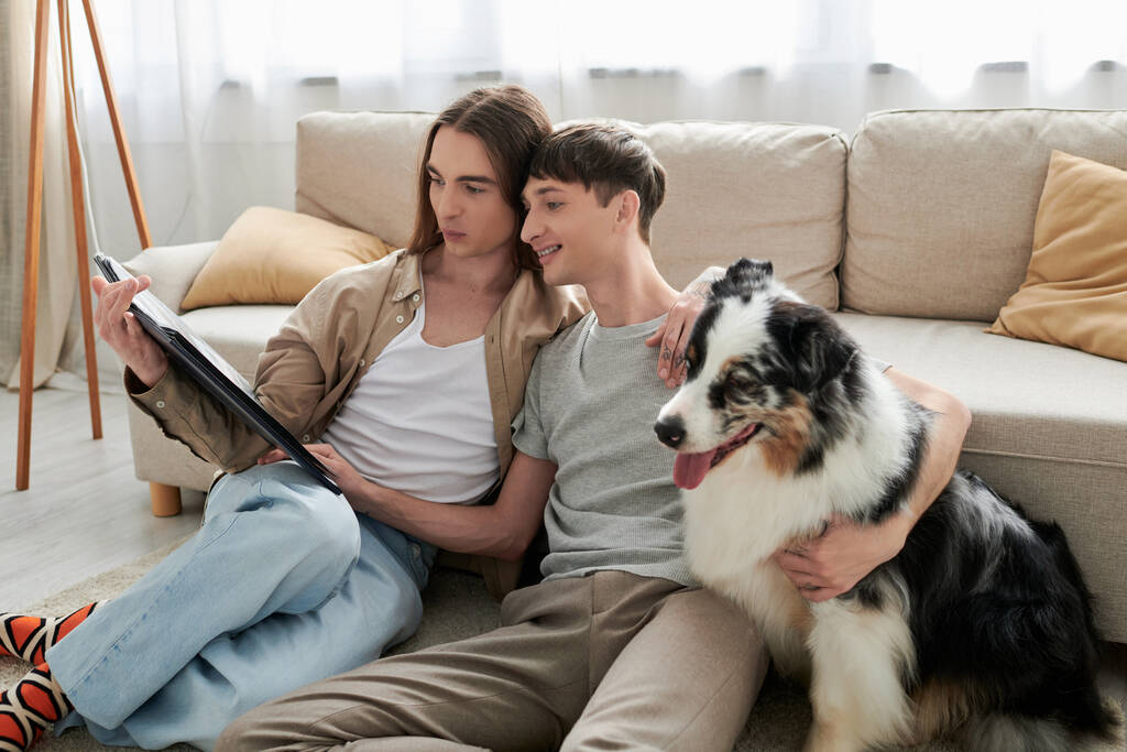 Lächelndes schwules Paar in lässiger Kleidung und Socken beim Betrachten eines geöffneten Fotoalbums neben pelzigem australischen Schäferhund und bequemer Couch im heimischen Wohnzimmer  - Foto, Bild