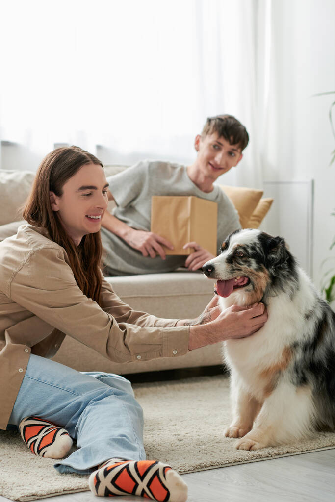 fröhlicher schwuler Mann mit langen Haaren kuschelt australischen Schäferhund, während sein Freund in lässiger Kleidung auf der Couch ruht und Buch im modernen Wohnzimmer hält  - Foto, Bild