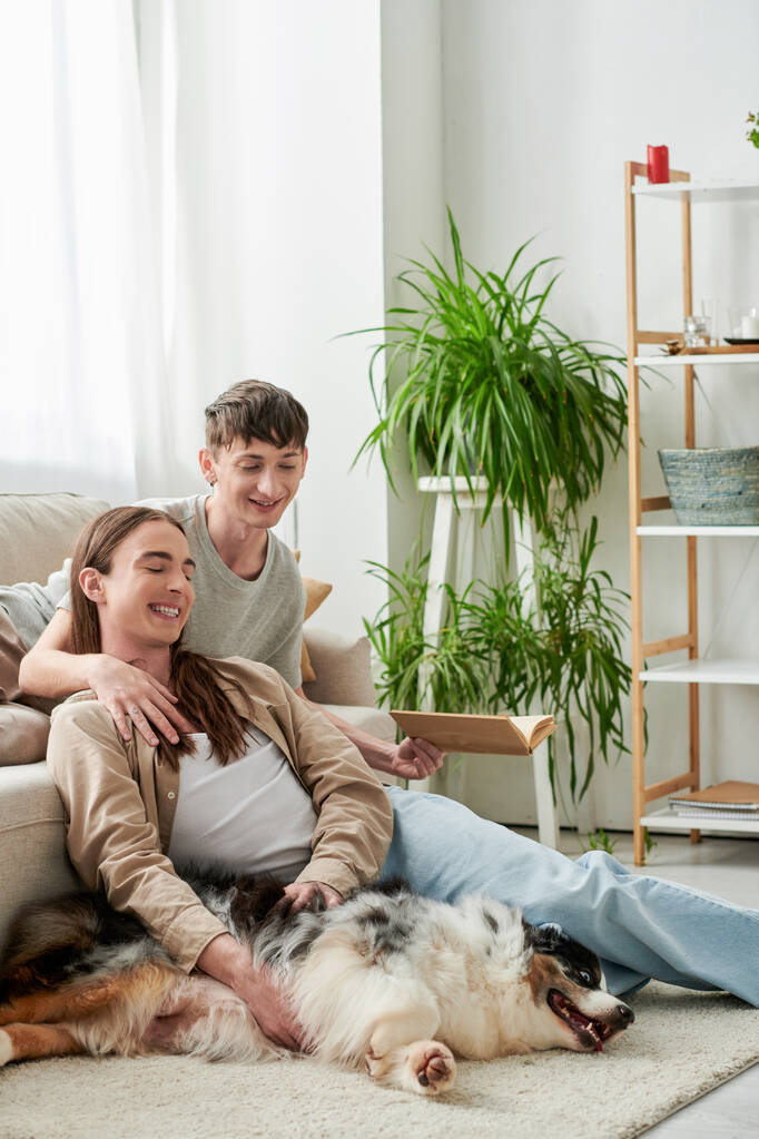 gai gay homme tenant livre et reposant sur canapé confortable tout en embrassant son petit ami avec les cheveux longs assis sur le tapis à côté de chien berger australien dans l'appartement moderne  - Photo, image