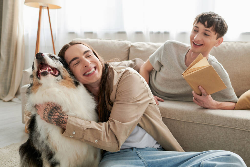 szczęśliwy gej człowiek z tatuaż uśmiech podczas przytulanie australijski owczarek pies obok wesoły gej człowiek gospodarstwa książki i spoczywa na wygodne kanapa w salon  - Zdjęcie, obraz