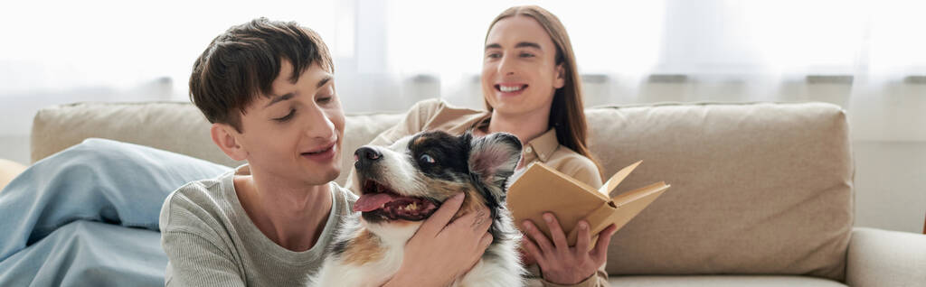 happy young gay man with long hair holding book and resting on comfortable sofa next to his boyfriend cuddling Australian shepherd dog in modern apartment, banner  - Photo, Image