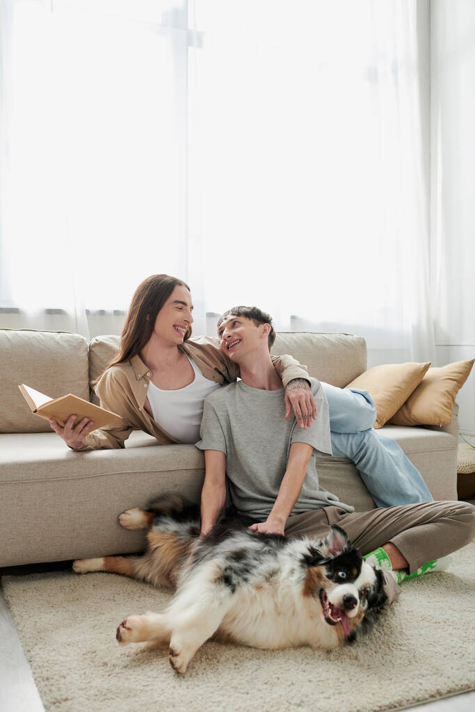 alegre gay homem sentado no tapete e abraçando australiano pastor cão e olhando para feliz parceiro com longo cabelo segurando livro no moderno sala de estar  - Foto, Imagem