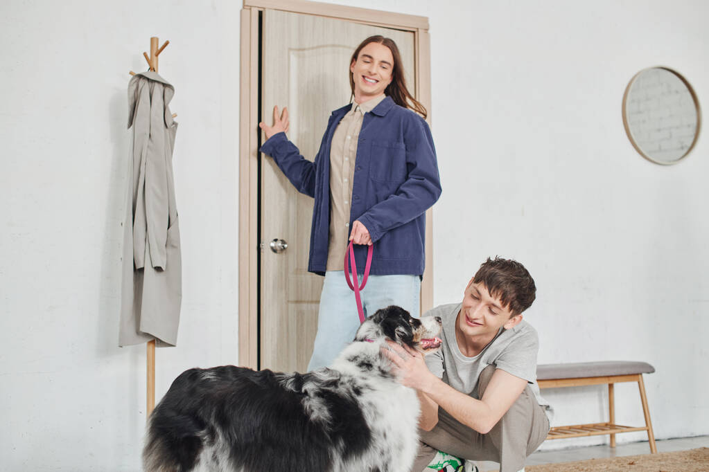 feliz gay homem sorrindo enquanto abraçando adorável australiano pastor cão ao lado de alegre namorado com longo cabelo segurando trela enquanto de pé perto da porta no moderno corredor  - Foto, Imagem