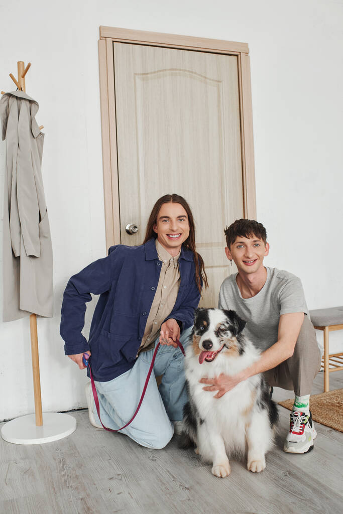 Mutlu eşcinsel çift, gündelik giysiler içinde gülümserken birlikte diz çöküp Avustralya çoban köpeğine sarılırken modern apartmanın koridorunda kapı ve askılık yanında.  - Fotoğraf, Görsel