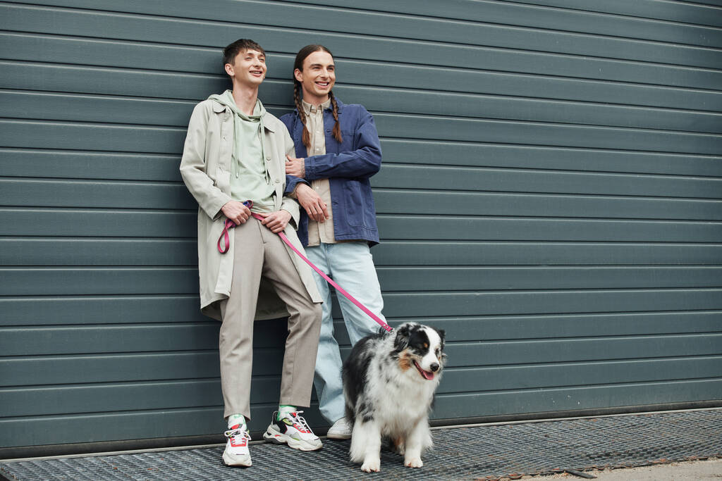 allegro gay uomo tenendo guinzaglio di australiano pastore cane e in piedi accanto a sorridente fidanzato con trecce vicino garage porta al di fuori su urbano strada - Foto, immagini