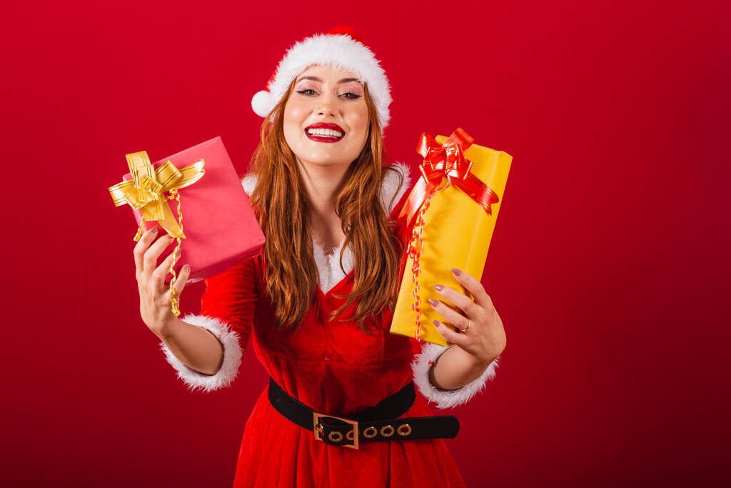 όμορφη βραζιλιάνα κοκκινομάλλα, ντυμένη με χριστουγεννιάτικα ρούχα, ο Άγιος Βασίλης. δίνοντας δύο δώρα στην κάμερα. - Φωτογραφία, εικόνα