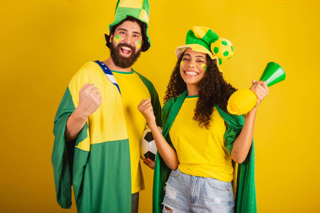 Ein paar brasilianische Fußballfans, gekleidet in den Farben der Nation, schwarze Frau, kaukasischer Mann. mit Flagge und Zubehör - Foto, Bild
