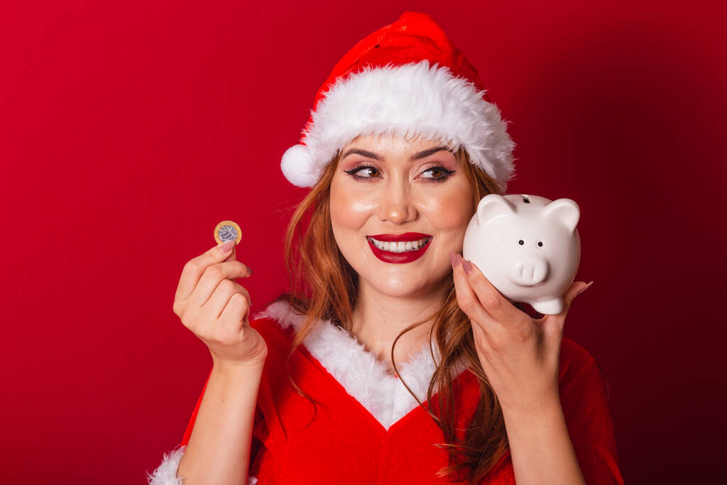 piękna brazylijska rudowłosa kobieta, ubrana w świąteczne ubrania, Święty Mikołaj. trzymanie skarbonki i monety. - Zdjęcie, obraz