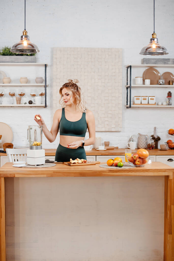 Vertikální pohled na blonďatou fitness ženu v zeleném sportovním oblečení stojící v kuchyni a dělá čerstvý pomerančový džus. Koncepce zdravé výživy a životního stylu - Fotografie, Obrázek