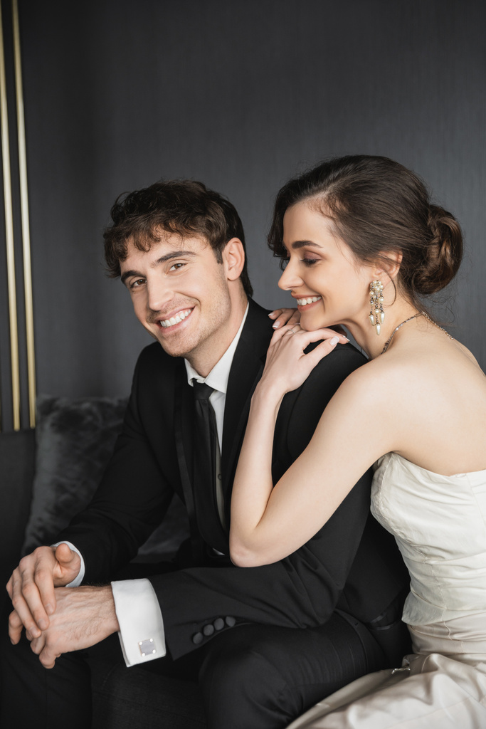 ritratto di sposa affascinante in abito da sposa bianco sorridente vicino bello sposo in abito nero guardando la fotocamera mentre seduto in camera d'albergo, sposi felici - Foto, immagini