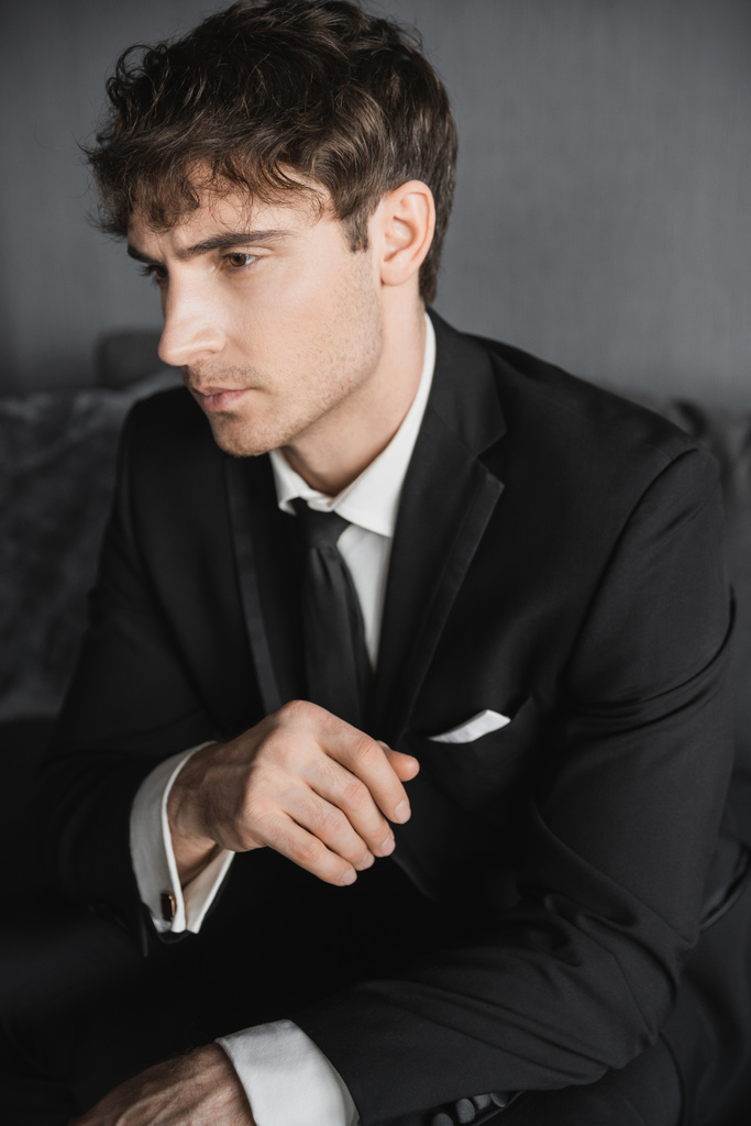 portrét dobře vypadajícího mladého ženicha v černém obleku s bílou košilí a kravatou sedí na pohodlné tmavě šedé pohovce v moderním hotelovém pokoji v svatební den - Fotografie, Obrázek
