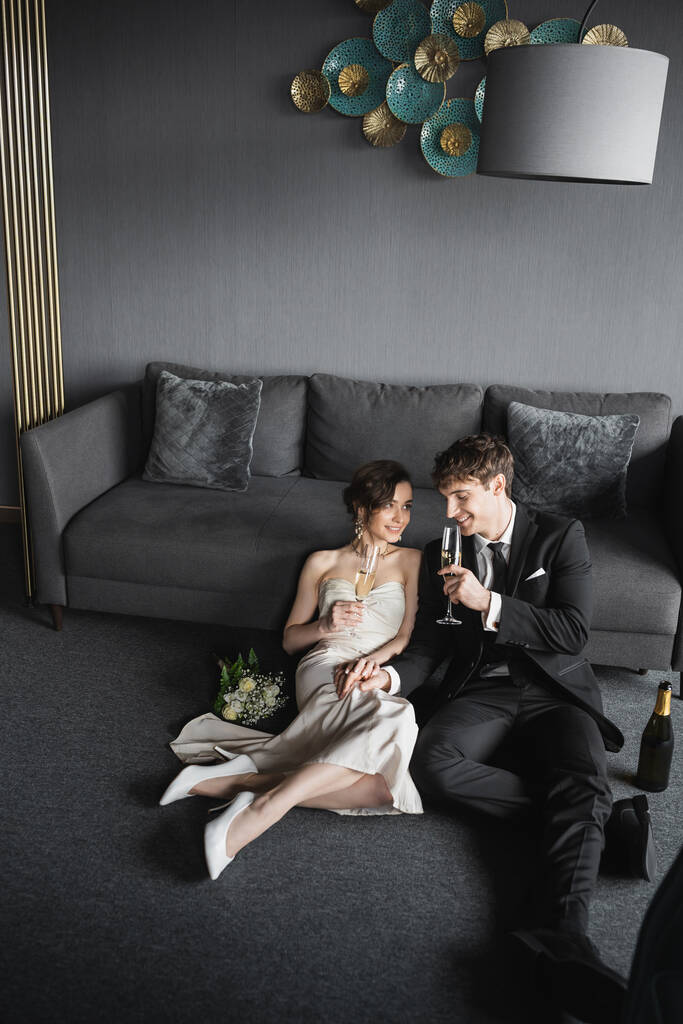χαρούμενη νύφη με λευκό φόρεμα και γαμπρός με μαύρο κοστούμι κρατώντας ποτήρια σαμπάνιας ενώ γιορτάζουν το γάμο τους κοντά σε νυφικό μπουκέτο και καναπέ μετά το γάμο στο δωμάτιο του ξενοδοχείου  - Φωτογραφία, εικόνα