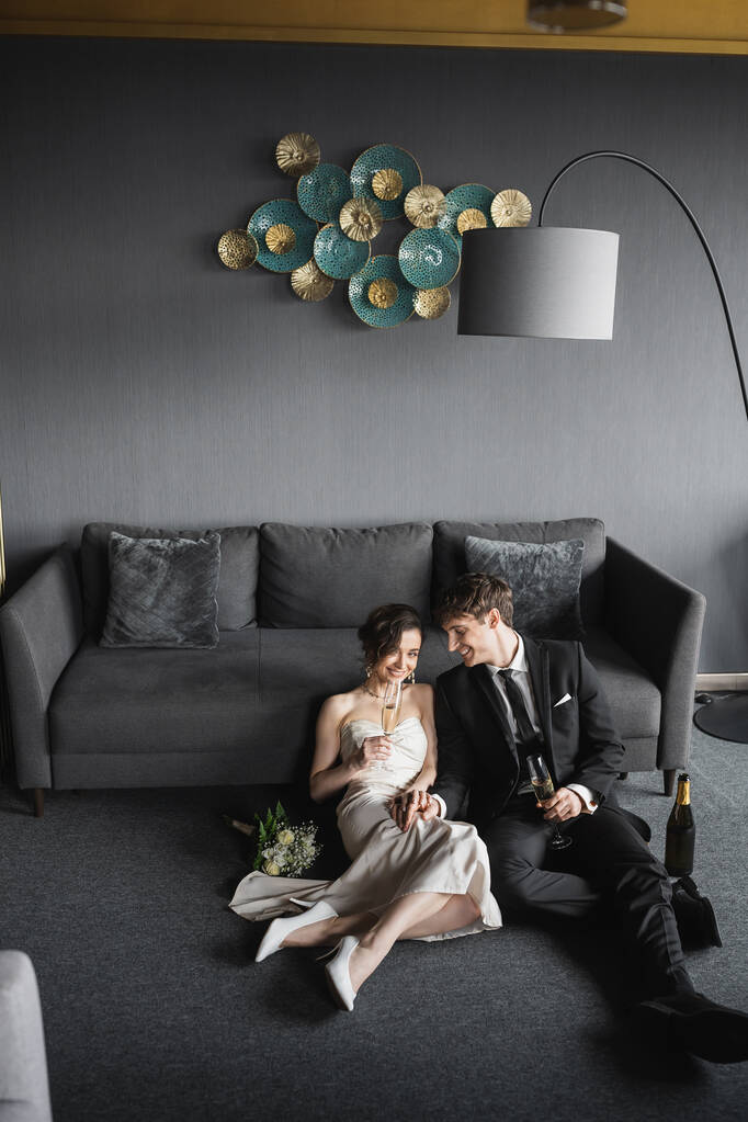 fröhliche Braut im Brautkleid und Bräutigam im schwarzen Anzug trinken Champagner, während sie ihre Hochzeit in der Nähe von Brautstrauß, Couch und Stehlampe im Hotelzimmer feiern  - Foto, Bild